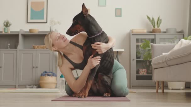 身穿运动服的健康白人妇女 坐在客厅的地板上 拥抱和亲吻成年的多伯曼犬 — 图库视频影像