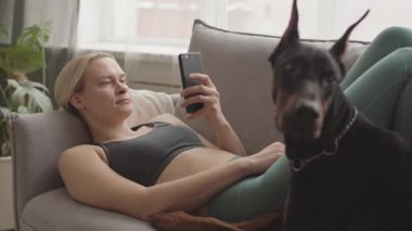 Sarışın, beyaz kadın idman kıyafetleri giyiyor, akıllı telefon kullanıyor, siyah köpeğiyle kanepede yatıyor.