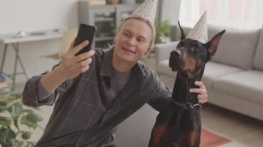 Kafkasyalı genç bir kadın ve siyah bir Doberman köpeği parti şapkası takıp evde selfie çekiyorlar.
