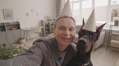 Beyaz kadın ve parti şapkası takan, evde selfie çeken, gülümseyen ve evcil hayvanı öpen siyah Doberman köpeğinin el bilgisayarı görüntüsü.