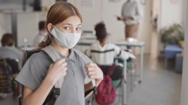 Sınıftaki arkadaşları arka planda bulanık öğretmenini dinlerken gri okul elbisesi ve yüz maskesi takmış, sırt çantasıyla kameraya poz veren, gülümseyen bir genç kızın orta derece yavaşlığı.