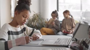 Modern sınıftaki sınıf arkadaşları bulanık arka planda sohbet ederken bilgisayarın başında oturmuş Afrika kökenli 5 ya da 6 ilkokul öğrencisi resimlerini çiziyor.