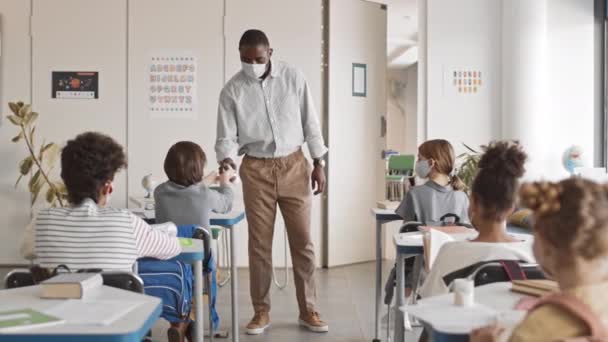 负责任的非洲裔美国教师身穿聪明的便衣 戴着面具 手握消毒手套 坐在办公桌前 手里拿着清洁剂 慢吞吞地走在教室的一排教室里 — 图库视频影像
