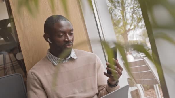 照片中像非洲裔美国人一样自信的男人坐在靠近窗户的咖啡桌旁 通过智能手机无线耳机聊天 — 图库视频影像