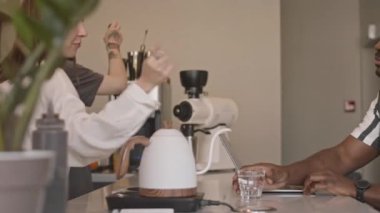 Kafedeki barda dizüstü bilgisayarlı Afro-Amerikalı bir adamın orta boy fotoğrafı. Barista taze kahve yapıyor ve garson da bir bardak su servis ediyor.