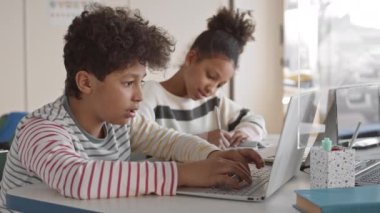 Orta boy Afro-Amerikan öğrencinin sınıfta dizüstü bilgisayarla yazarken kız kardeşiyle yazışırken, deftere not alırken çekilmiş bir fotoğrafı.