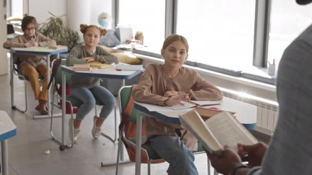 手を上げと現代の教室で彼らの机のそばに座っている彼らの前に立っている先生の質問に答える準備ができて彼らの前に本 — ストック動画
