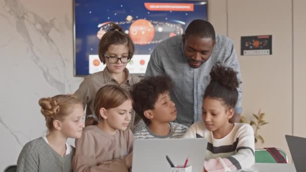 多民族の子供たちとその若い男性の中低速アフリカ系アメリカ人の先生は サイエンスクラス中にノートパソコンの画面を一緒に見て楽しんでいます — ストック動画