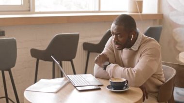 Kulaklıklı, modern Afro-Amerikan iş adamının laptopta iş ortaklarıyla kafeteryada otururken görüntüsü.