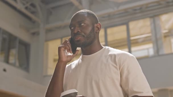 穿着T恤衫 拿着一杯咖啡 用智能手机聊天 站在现代建筑内部的年轻非洲裔美国人的提神镜头 — 图库视频影像