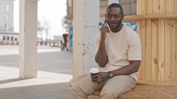 中ショットの陽気な若いアフリカ系アメリカ人男性ですカジュアルウェアで屋外の木製のベンチに座ってコーヒー電話を作ります — ストック動画