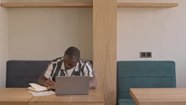 非洲裔美国年轻自由撰稿人穿着便服在咖啡桌边打趣 笔记本电脑在抄袭本上记笔记的中景照片 — 图库视频影像