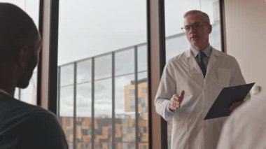 Tıp konferansı ya da seminer sırasında Afro Amerikalı meslektaşımla konuşurken gözlüklü ve laboratuvar önlüklü olgun bir doktorun orta yavaş konuşması.