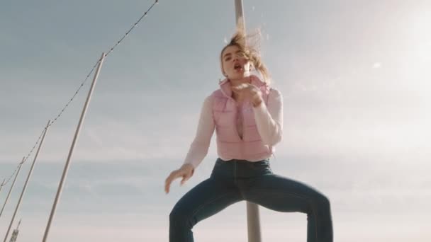 穿着休闲裤和宽松夹克 身披蓝天前卫柱子跳舞的低角度中等身材的活跃的白人年轻女子 — 图库视频影像