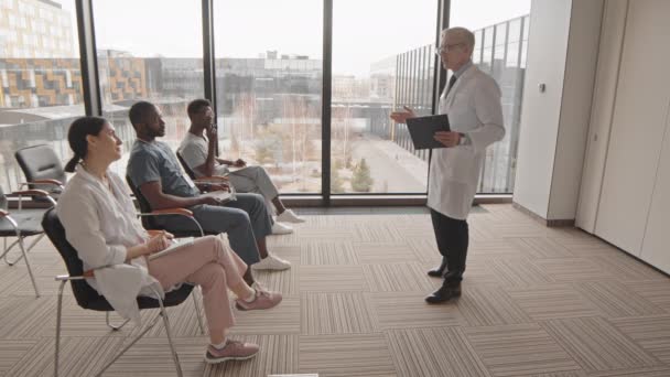在明亮的现代大厅举行的医学会议或研讨会上 穿着实验室外套 手持剪贴板 带着自信的成熟医生在同事面前发言时的侧视慢度 — 图库视频影像