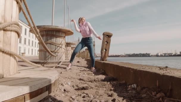 穿着休闲裤和宽松夹克的活跃的白人年轻女子的全景 在湖滨蓝天的前景下 在巨大的木制箱子边跳舞 — 图库视频影像