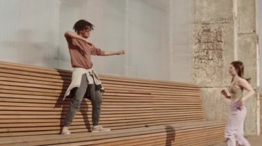 Genç enerjik Afrikalı adamın ahşap bankta dans etmesi, aktif beyaz bir kadının katılması.