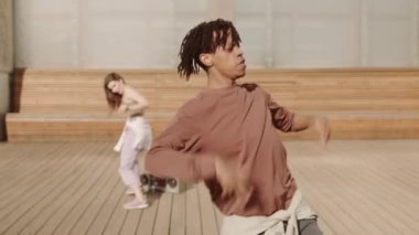 Sıcak ve güneşli bir günde kamerada çeşitli hareketler yapan Afrikalı genç erkek dansçının orta uzunluktaki görüntüsü arka planda dans eden bulanık kadın.