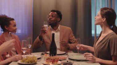 Orta yaşlı, mutlu, Afro-Amerikalı bir adamın yemek masasında birlikte oturan arkadaşlarıyla kadeh tokuşturması.