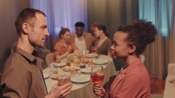 友人がパーティー中に背景にぼやけて夕食のテーブルに座っている間 お互いの話の前にワインのグラスが立っている若い多民族のカップルのサイドビュー中低速 — ストック動画