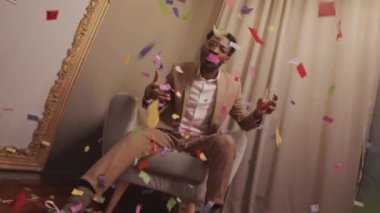 Elinde şampanya flütü tutan, kameraya el kol hareketi yapan lüks bir dairede oturmuş konfetiler düşerken kameraya el ele tutuşan Afro-Amerikan genç adam.