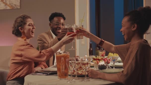 中低速の二人の美しい若い女性と彼らの陽気なアフリカ系アメリカ人男性友人クリンクカクテルグラスホームディナーパーティー中に特別な機会に — ストック動画