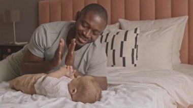 Orta uzunlukta bir Afrikalı baba çocuğuna el çırpmayı öğretiyor, yatakta uzanıyor, gülümsüyor ve konuşuyor.