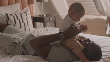 Orta boy mutlu, genç Afrikalı bir adam yatakta uzanmış, yeni yürümeye başlamış oğlunun ellerini çırpıyor, babasının gülümsemesi ve çocuğuyla konuşması.