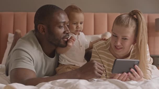 Στήθος Πολυεθνικών Γονιών Ξαπλωμένοι Στο Κρεβάτι Χρησιμοποιώντας Smartphone Μιλώντας Μικρός — Αρχείο Βίντεο