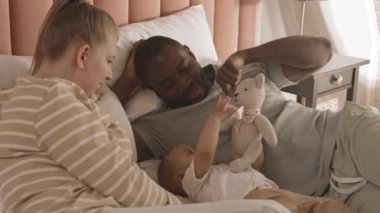 Yatakta yatıp oyuncak ayılarla oynayan melez bebeklerini izleyen mutlu anne babaların yüksek açılı görüntüsü.