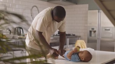 Orta uzunlukta bekar bir Afrikalı erkek ebeveyn telefonda konuşuyor ve küçük oğlunu mutfak masasında bebek bezi ve pantolonla giydiriyor.