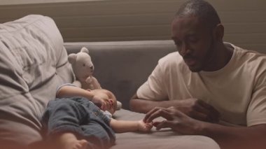 Orta boy, kanepede oturan, kanepede uyuyan huzurlu melez bir bebeğin minik ellerine dokunan Afrikalı bir adam.