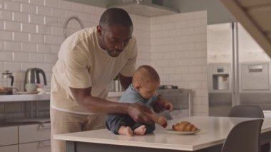 Orta uzunlukta bir Afrikalı genç adam mutfakta duruyor, bir yaşındaki oğlunun masada oturup çilekle oynamasını izliyor ve ona yardım ediyor.
