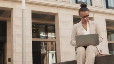 Şehir merkezindeki modern ofis binasının yanındaki beton bankta otururken dizüstü bilgisayarda çalışan, bej takım elbiseli, orta yaşlı, çekici, melez iş kadını.