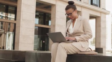 Modern ofis binasının dışındaki beton bankta otururken bilgisayarla yazı yazma ve iş arkadaşlarıyla görüntülü konuşma üzerine çalışan akıllı gündelik kıyafetli genç iş kadınlarının orta derecede yavaşlaması.