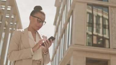 Düşük açılı orta açılı orta açı çekim. Yüksek topuz ve bej takım elbiseli, kendinden emin iş kadını güneşli bir günde şehirde yürürken akıllı telefon kullanıyor.