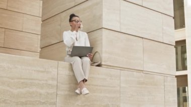 Modern ofis binasının önünde dizüstü bilgisayarda çalışan ve bacakları sarkık halde oturan güncel iş kadınlarının yavaş çekim yaptığı geniş açılı bir çekim.