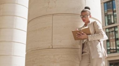 Düşük açılı geniş açılı çekim. Gözlüklü ve kırmızı rujlu düşünceli genç iş kadını şehir merkezindeki planlayıcısında durup büyük sütunlara yaslanıyor.