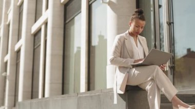 Gündüz güneşli bir günde modern ofis binasının yanındaki beton bankta oturan genç ve çekici iş kadınının laptopta çalışmasını yavaş çekim.