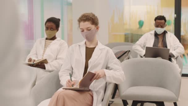 フェイスマスクと白いコートを着た3人の若い多民族医学の学生の中低速セミナー中に教授の質問に答えるために手を上げる — ストック動画