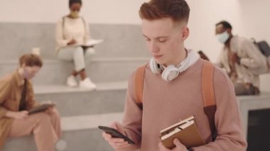 Modern üniversite salonundaki maskeli sınıf arkadaşları arka planda otururken, akıllı telefonlu genç erkek öğrenci portresi ve günlük kıyafetleri kameraya poz veriyor.