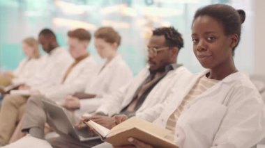 Gülümseyen Afro-Amerikan tıp öğrencisinin fotoğraf makinesine notlar alıp modern oditoryumdaki sınıf arkadaşlarıyla aynı sırada oturarak konferans verirken orta boy yavaş portresi.