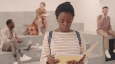 Üniversitede kameraya poz veren Afro-Amerikalı mutlu genç bir kızın orta yavaş portresi.