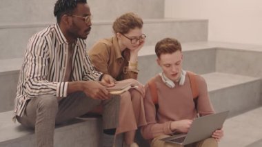 Projeye hazırlanan üç genç üniversite öğrencisinin orta derecede yavaşlaması. Detayları tartışıyorlar ve modern çalışma alanında merdivenlerde otururken dizüstü bilgisayar kullanıyorlar.