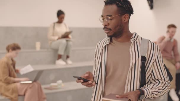 中低速肖像画の普及した若い男性アフリカ系アメリカ人大学生カジュアルな服や眼鏡カメラのポーズでスマートフォンが室内に立っている現代のコワーキングスペース — ストック動画