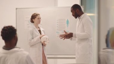 Orta boy Afro-Amerikan Anatomi Profesörü sınıfta materyalleri açıklarken bayan tıp öğrencisi onu elinde insan beyni mankeni tutarken dinliyor.