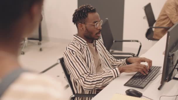 プログラム開発を勉強するコンピュータの前に座っている若いアフリカ系アメリカ人の男性の肩の上中低速同級生と会話をしながら — ストック動画