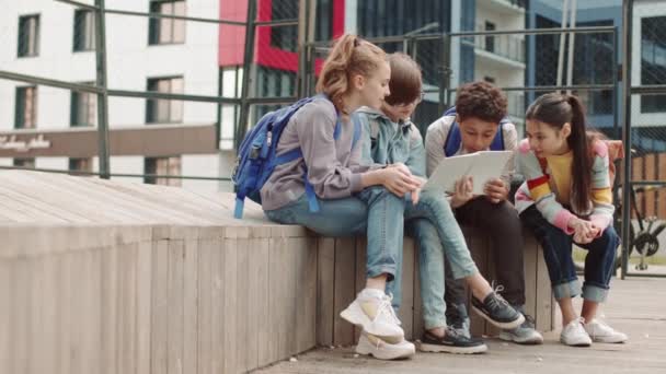 コピーブックを見ながら夏のチャットで遊び場でベンチに座っている未就学児のグループのスローショット — ストック動画