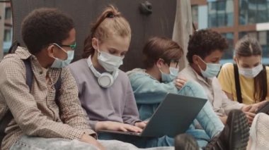 Ev ödevi yaparken bilgisayar ve dijital tablet kullanarak okuldan sonra dışarıda maske takan çok ırklı bir grup gencin yavaş çekimleri.