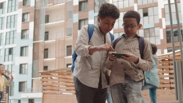 两名男生在智能手机上玩游戏 而他们的同学在操场上玩球的低角度慢镜头 — 图库视频影像